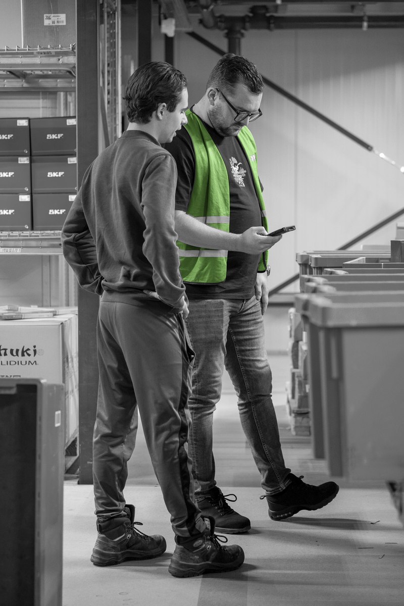 Twee medewerkers overleggen met een handterminal bij de magazijnstellingen.